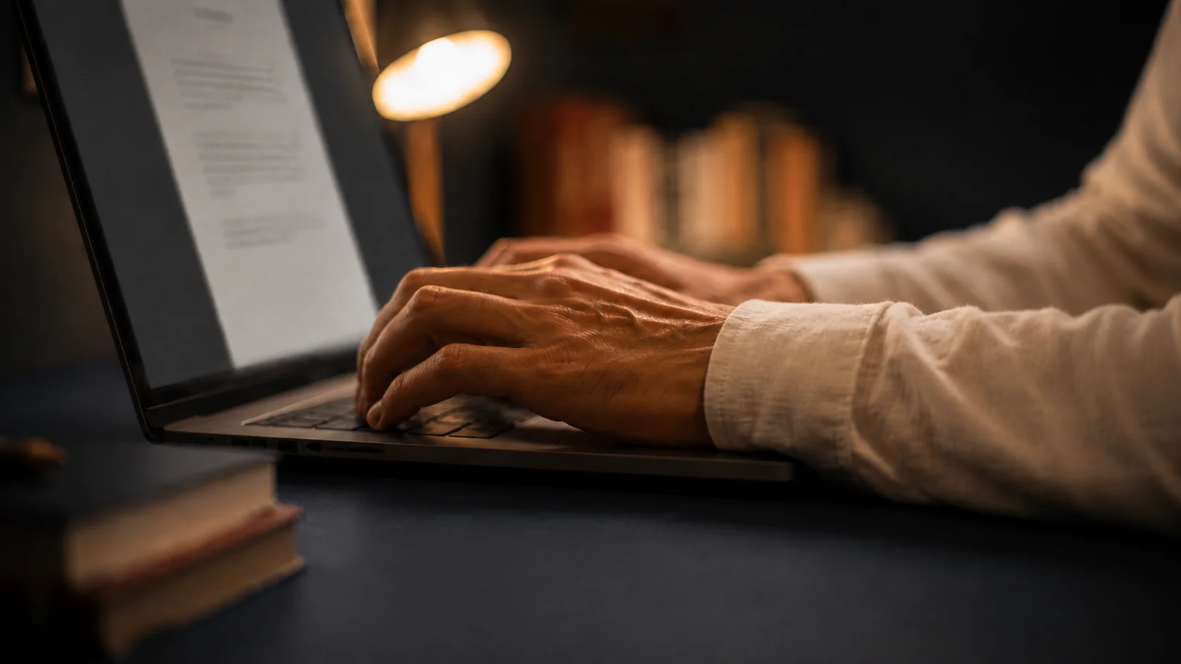 Hände tippen auf einem Laptop in warmem Licht; Schirm, Lampe und Bücher bleiben sanft unscharf.